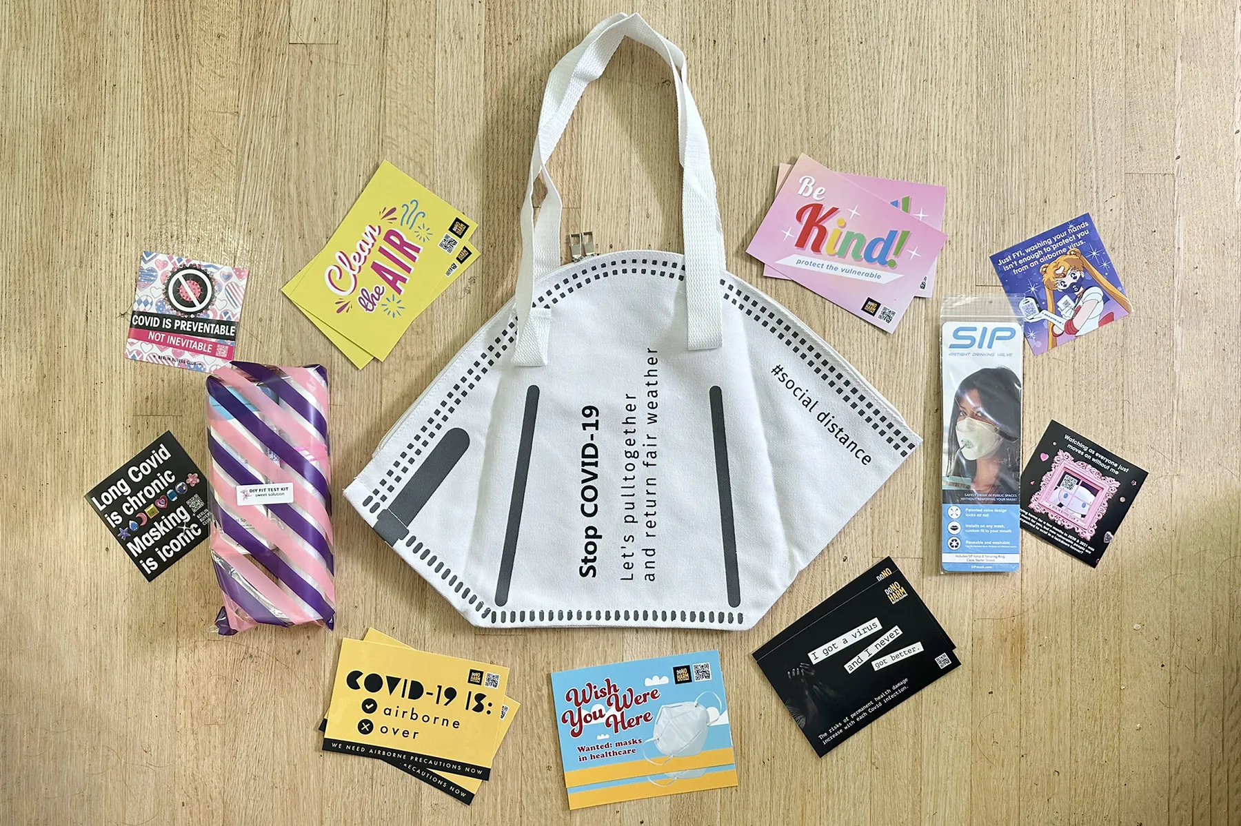 Around a white N95 shaped tote bag are colourful DoNoHarm BC postcards and Berlin Buyers Club stickers, plus a sip valve and a DIY fit test kit in a colourful goodie bag